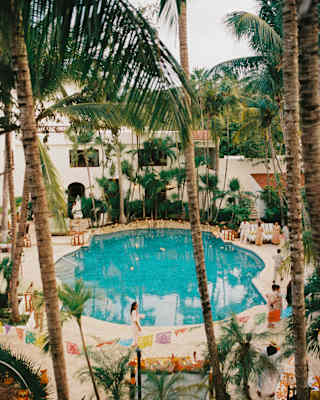 Guests chat at the edge of the tropical pool where bright Papel Picado bunting is strung between the palms, seen from above.