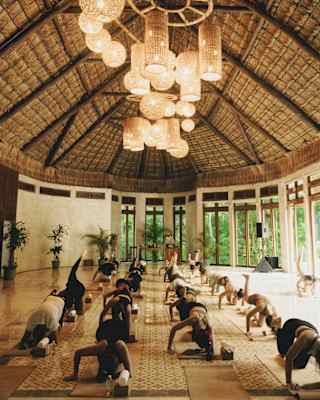 In a high-ceilinged palapa lit by a cluster of wicker pendant lights, twenty guests practise yoga on mats, arranged in four rows.