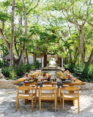 A wooden table laid for six, with black and white napkins and flower displays, sits in dappled shade by Ranas Fountain.