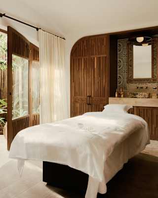 A white bed bathes in light from the large windows of a spa room with wood panels, towel rack, marble sink and garden views.