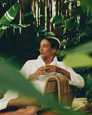 A woman in a white robe relaxes on a cane chair with a drink in the Maroma Spa by Guerlain courtyard, viewed through foliage.