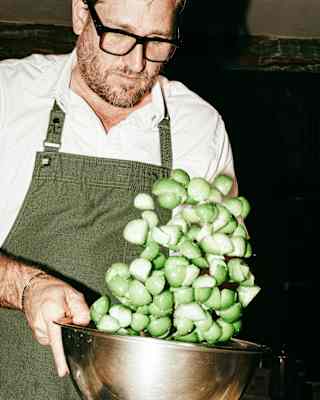 Chef Curtis Stone, with a short beard and dark-rimmed glasses, tosses chopped green tomatoes in a steel bowl, seen close-up.