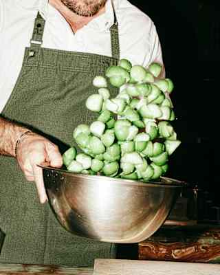 A person in a green apron tosses a metal bowl filled with fresh green Brussels sprouts, causing some to rise into the air above the bowl. The person's face is partially out of the frame.