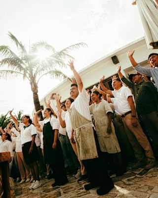 A group of people standing outdoors, raising their right arms in unison under a bright sky near a palm tree; some wear aprons, suggesting a team or staff gathering.