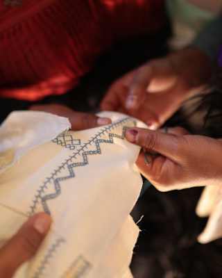 An embroiderer stitches track and zigzag patterns on to white cotton as another craftsperson holds it taut, seen in closeup.