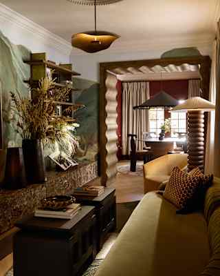 View through a living room with green sofa, coffee table and wheaten flowers to a dining area, divided by a carved doorframe.