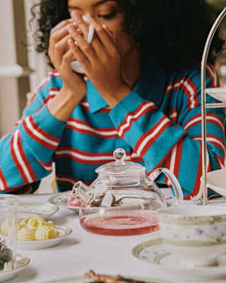 A dark-haired female guest in a blue shirt with red and white stripes drinks from her cup using both hands at afternoon tea.
