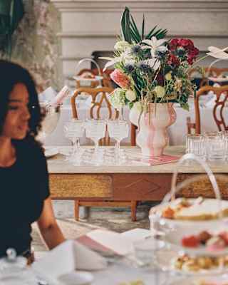 A white ornate metal table set for tea outdoors, with a teapot, teacup, saucer, pink macarons on a plate, and folded white serviettes. The background shows green bushes and pink flowers.