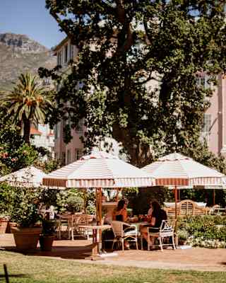 Four guests dine in the shade of large coral and white-striped parasols on a flower-filled terrace, with a mountain backdrop.