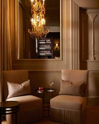 A cosy lounge area with two beige chairs, a small round table holding two drinks, and a chandelier hanging above; warm lighting and elegant décor create a sophisticated atmosphere.