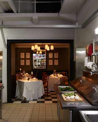 View across the working kitchen to a miniature dining room with checked tiles and Cortez chandelier which glows above two tables.