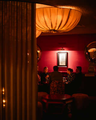 A table of guests chat below warm orange cloth pendant lamps in the moody interior of The Red Room, seen through a doorway.