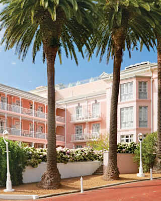 A large, elegant pink hotel with white balconies is framed by tall palm trees and greenery on a sunny day, with a clear blue sky in the background.