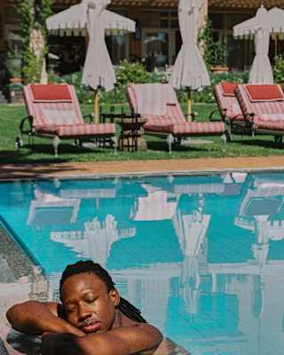 A man rests at the side of the pool while behind him loungers and parasols drop their reflections on the clear blue water.