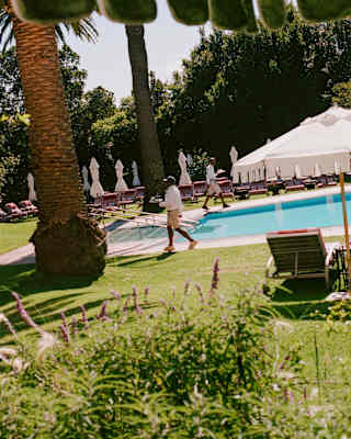 Staff walk either side of the pool in view from beneath a parasol, capturing gardens, rows of loungers and a white marquee.