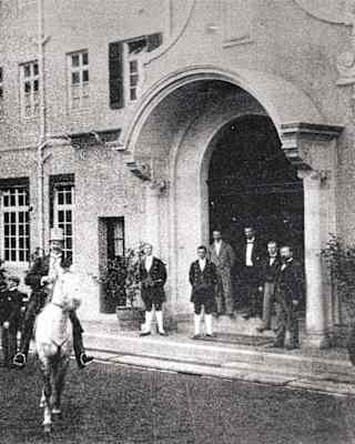 A black-and-white photo shows two people on horseback in front of a building entrance, whilst a group of people stands by the doorway, dressed in early 20th-century clothing.