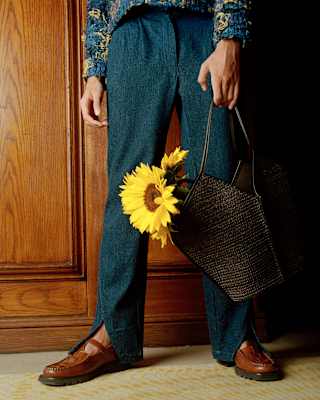 A man in dark blue trousers with splits revealing brown T-bar loafers, carries a bag of sunflowers, seen from the waist down.