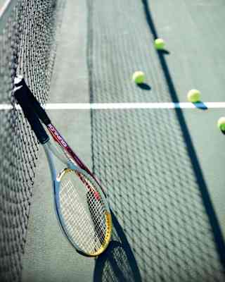 Two tennis rackets lean against the net, which casts a shadow on the green court, where balls have been left during a break.