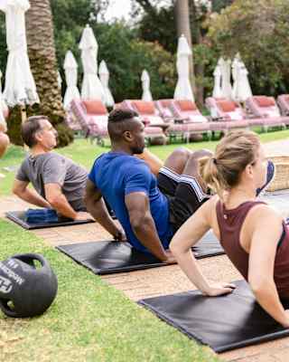 Two men and a woman perform core exercises on black mats, seen in a side-view that captures garden loungers on lawn behind.