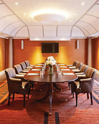 Looking down a long table with glasses, clipboards and four chairs either side in a bright room with warm lights and a tv.