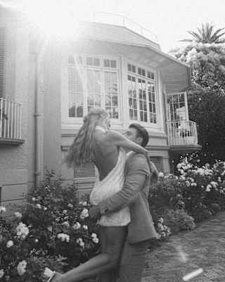 In a flowering garden, a suited man lifts a woman with long blonde hair and a low-back white dress, captured in monochrome.