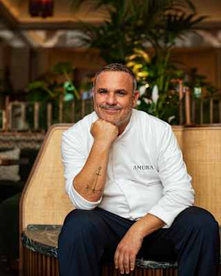 Chef Ángel León smiles to camera as he sits in Amura in a branded white top and dark trousers, resting his chin on his fist.