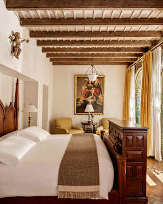 Sun spills through two arched windows into a bedroom with stone and painted walls, viewed over a bed below a beamed ceiling.