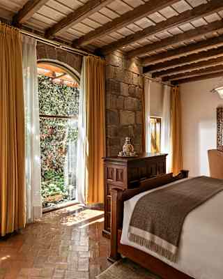 Sunlit bedroom with large arched windows, yellow curtains, and a rustic wooden ceiling. The room has a bed, a wooden desk with a mirror, and looks out onto a lush, green garden with ivy-covered walls.