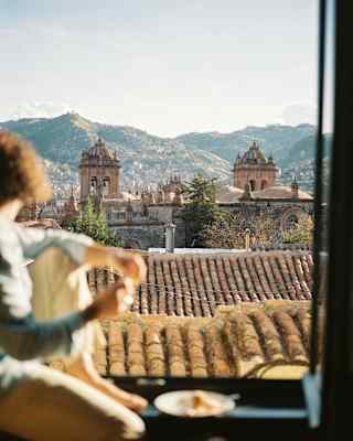 A woman in soft-focus foreground sits on the sill of an open window gazing towards Cusco Cathedral and the mountains beyond.