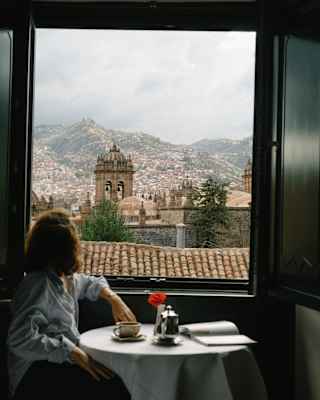A woman sits at a table with coffee gazing out of her suite window at the terracotta roofscape of Cusco beneath a moody sky.