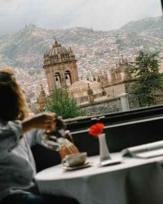 View of Cusco's roofscape, basilica bell tower and mountains, seen from a window where a woman pours coffee in soft-focus.