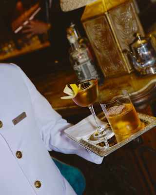 A waiter carries two old-fashioned style cocktails on a tray, one with ice, the other with lemon rind garnish, seen close-up.