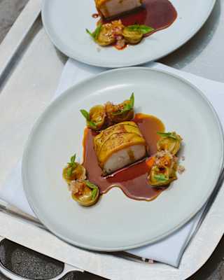 A plated gourmet dish featuring a rolled piece of meat with a golden exterior, served with sauce, garnished vegetables, and small green tortellini on a white plate atop a silver tray.