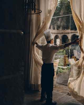 A chef in a toque pushes open drapes at the glass entrance to Oqre, letting in light and courtyard views, seen from behind.