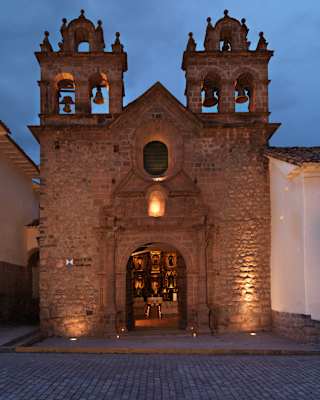 Front view of the ancient stone façade of Monasterio, with twin bell towers and a large open arched door, spot-lit at dusk.