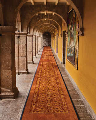 A long corridor with stone arches, a patterned red carpet, and a yellow wall adorned with framed artwork, creating a warm, historic atmosphere.