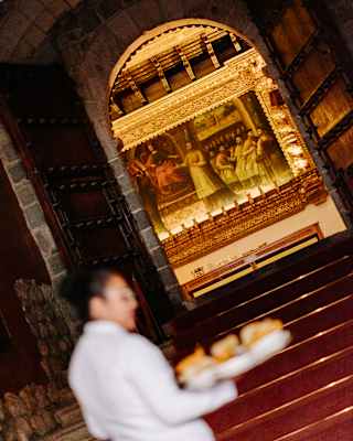 A person in white clothing carries a tray of food up red carpeted stairs towards a golden, ornately decorated altar or shrine inside a dimly lit building.