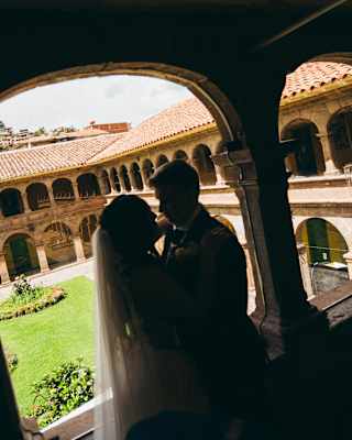 A bride and groom share a moment in shadow under an archway, with a sunny courtyard and historic stone building visible in the background.