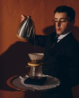 A barista creates pour-over coffee, tipping hot water into a filter over a glass jug, photographed against a red wall.