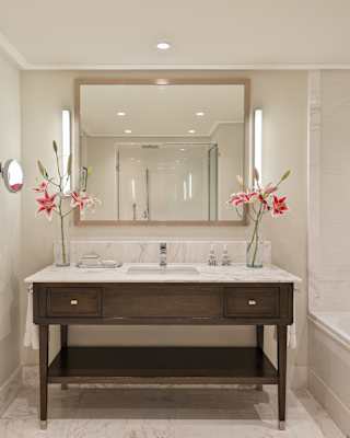 Two vases of pink Stargazers adorn the marble sink top of a dark-wood unit in a bathroom of bright white paint and tiles.