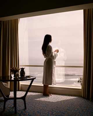 A woman with long dark hair stands in a robe by a huge suite window, holding a coffee and gazing out at the ocean below.