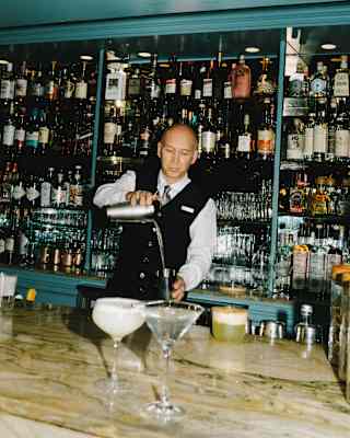 A barman pours a Martini from a shaker at a marble bar top alongside a ready-made pisco sour, with bottle shelves behind.
