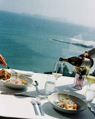 A guest dines as white wine is poured at a table for two at The Observatory Restaurant, with high views over the blue sea.
