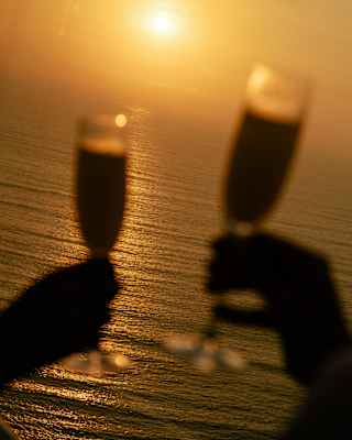 In soft-focus foreground, two hands raise Champagne glasses against the backdrop of an ocean tinted by a golden sunset.