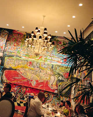Angled image of two tables of guests as they dine beneath a chandelier and the large vivid murals of artist Mateo Liébana.