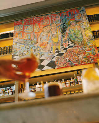 Low-angle view of a bright, busy Mateo Liébana artwork above Tragaluz bar, seen past a hand holding a drink in soft-focus.