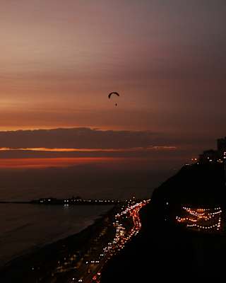 The hand-glider silhouetted against a crimson sunset sky soars above the trails of traffic lights along the Lima coastline.