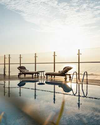 Two half-reclined sun-loungers face each other over a small table by the glass terrace barrier, reflected in the still pool.