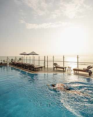 A man swims in the rooftop pool, in a view to the opposite side where eight loungers face away, towards the glowing ocean.