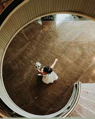 A dark-haired bride with a white dress and bouquet walks across a tiled room, in a view from above, taken from a spiral staircase.